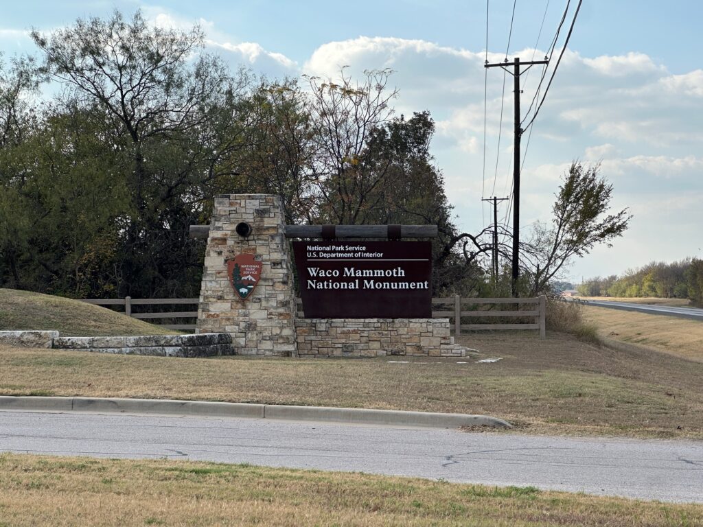 An outdoor interpretive panel titled “Discover the Waco Mammoths” stands among trees and fallen leaves along a paved path. The sign features sepia-toned images of two men in hard hats, a small photo of the dig shelter, and a map of North America showing mammoth sites. Text blocks explain the accidental discovery of the bones by local men and how the find became a major scientific site.