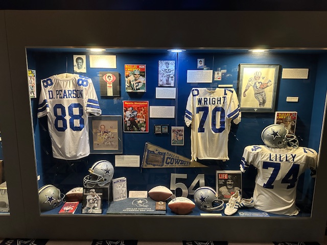 A glass display case at the Texas Sports Hall of Fame filled with Dallas Cowboys memorabilia: framed photos, helmets, footballs, and three iconic white and blue jerseys for players Drew Pearson (#88), Rayfield Wright (#70), and Bob Lilly (#74).