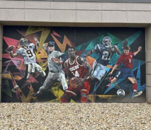A vivid outdoor mural covers a concrete wall, featuring five elite athletes from different sports frozen mid-action. A football quarterback, a baseball pitcher, a basketball player in a red “HOUSTON” jersey, an American football running back, and a soccer player in a red and blue kit all burst forward against a background of sharp, colourful geometric shapes. Pebbled ground fills the foreground, emphasizing that this is a large-scale exterior artwork.