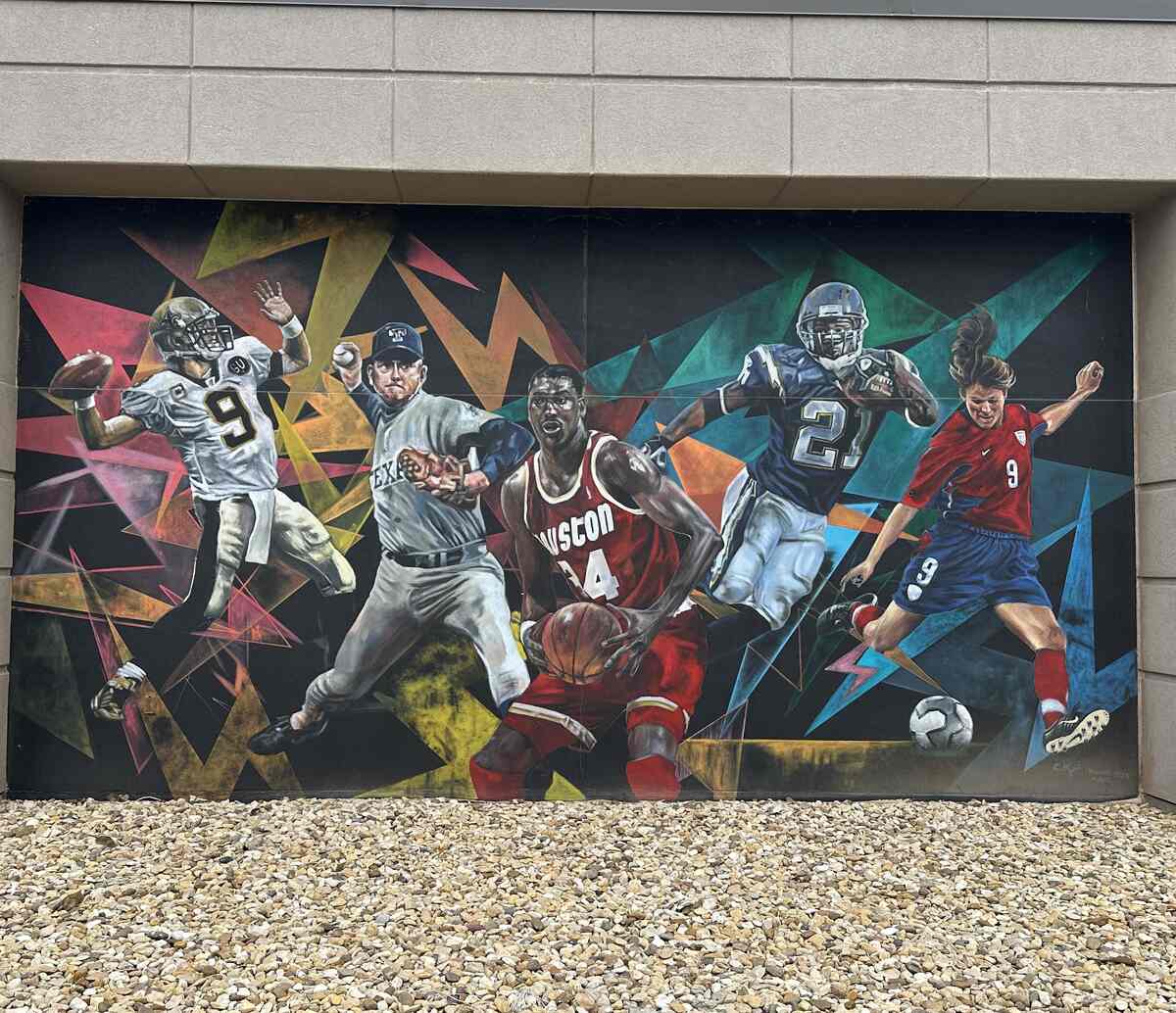 A vivid outdoor mural covers a concrete wall, featuring five elite athletes from different sports frozen mid-action. A football quarterback, a baseball pitcher, a basketball player in a red “HOUSTON” jersey, an American football running back, and a soccer player in a red and blue kit all burst forward against a background of sharp, colourful geometric shapes. Pebbled ground fills the foreground, emphasizing that this is a large-scale exterior artwork.