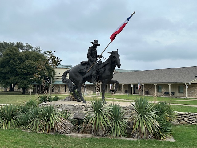 An outdoor bronze statue of a Texas Ranger on horseback holding the Texas flag aloft. The figure is framed by low stone walls and desert plants, with the Texas Ranger Hall of Fame and Museum buildings stretching out behind.