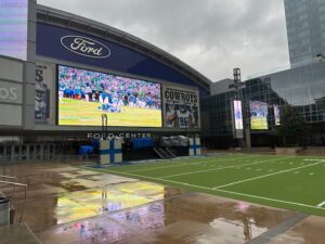 Wide view of the Ford Center at The Star in Frisco on a rainy day, with a giant video screen showing a Cowboys game above the entrance and reflections shimmering on the wet pavement beside the outdoor turf field.