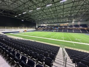 Wide interior view of the Ford Center stadium, showing rows of navy seats curving around a full football field beneath a high steel roof and bright stadium lights.