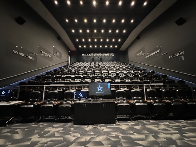 Dark, steeply tiered theatre filled with leather recliner seats facing a large screen, with words like “Work Ethic,” “Commitment,” “Accountability,” and “Passion” etched on the side walls.