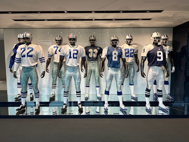 A row of mannequins dressed in Dallas Cowboys uniforms from different eras, showing the evolution of jerseys, helmets, and numbers across decades, all reflected on a glossy floor.