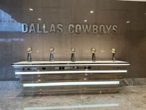 Five Vince Lombardi trophies displayed in a sleek marble and chrome lobby beneath large metallic letters spelling “DALLAS COWBOYS.”