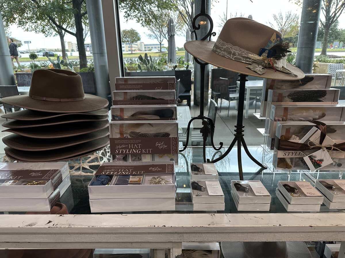 Display of wide-brimmed felt hats and “hat styling kit” boxes arranged on a rustic white table inside Heirloom Haul in Frisco, Texas, with custom hat bands and embellishments laid out in front and large windows showing a tree-lined walkway and lawn outside.