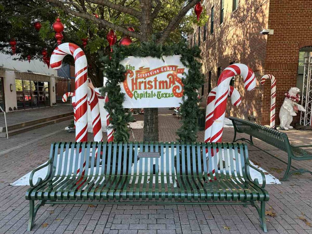 Daytime view of the same Christmas Capital of Texas sign, framed by candy cane poles, green benches, and brick pathways beneath leafy trees.