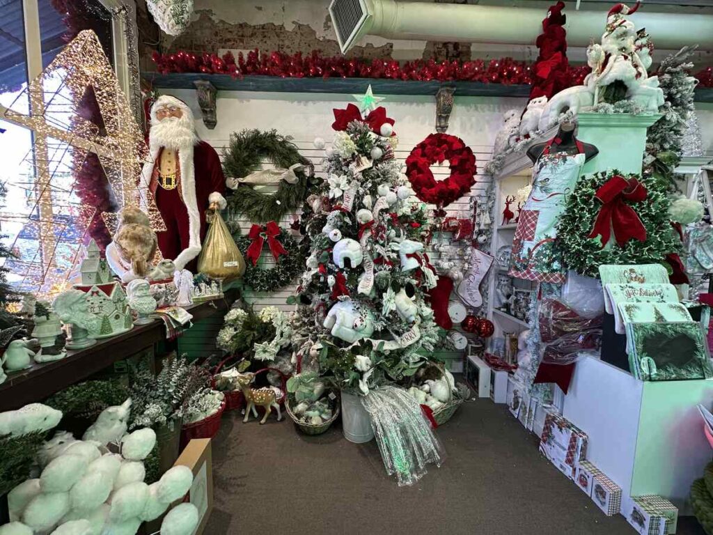 A packed holiday boutique filled with Christmas décor, from a fully decorated tree and wreaths to figurines, ornaments, and a life-sized Santa standing by the window.