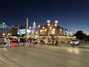 A festive corner building on Historic Main Street in Grapevine glows with layers of Christmas lights, wreaths, and window displays as people carrying pink shopping bags cross the street and cars roll past under a deep blue evening sky.