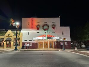 Nighttime shot of Grapevine’s Palace Theatre, its white façade trimmed with red tile, marquee lit up with classic Christmas movie titles, and wreaths hanging above the entrance.