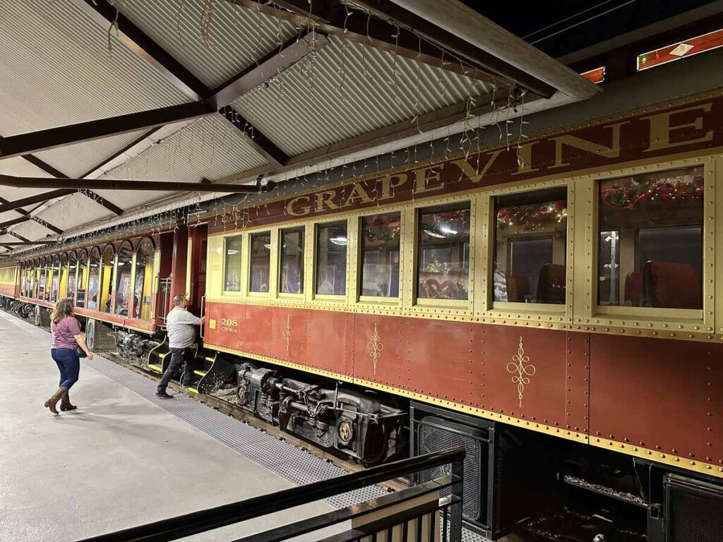 Travellers walk along the platform toward a red and gold Grapevine railcar, its windows trimmed with garlands and lights under a covered platform roof.