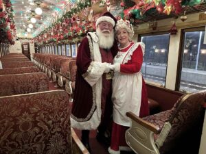Santa and Mrs. Claus stand arm in arm in the aisle of an ornately upholstered vintage coach, surrounded by red patterned seats and festive garlands overhead.