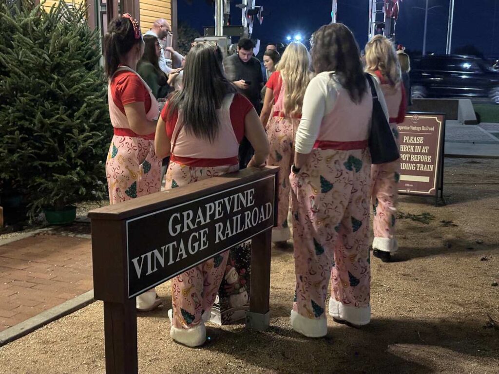 A group of women in matching pink Christmas pyjamas and furry slippers walk toward the Grapevine Vintage Railroad sign at the boarding area, with a Christmas tree nearby.