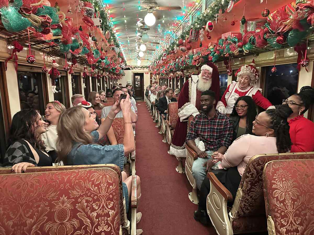 A lively vintage railcar packed with smiling passengers as Santa and Mrs. Claus stop to greet a group, with colourful garlands, bows, and lights decorating the entire ceiling.