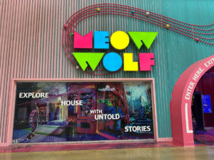 A vibrant Meow Wolf entrance at Grapevine Mills, with a teal and pink striped wall, a bold neon “MEOW WOLF” sign in bright colours, and a large window graphic inviting visitors to “Explore a house with untold stories.”
