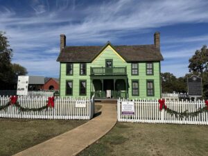 A bright green, two-storey historic farmhouse with a white picket fence trimmed in garlands and red bows, set under a blue sky at Nash Farm.