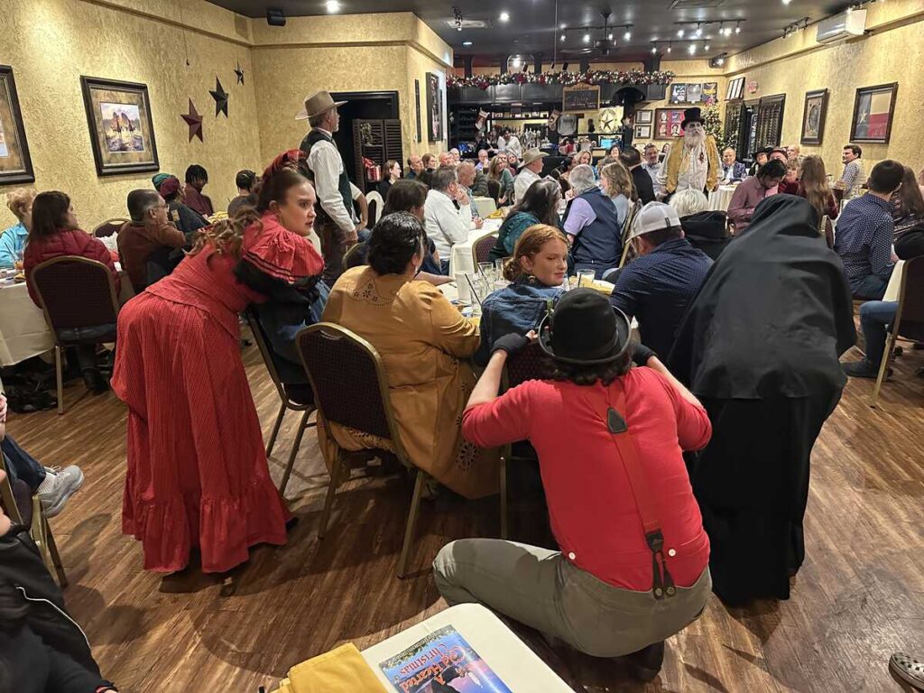 Guests seated at banquet tables inside a warm, gold-toned dining room while costumed actors in period Western attire move between tables during a performance.