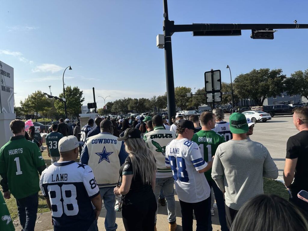 A crowd of fans in Cowboys blue and Eagles green jerseys walks along a sunny intersection outside the stadium, heading towards the entrances under a clear Texas sky.