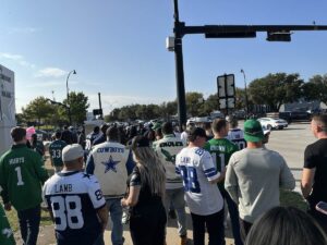 A crowd of fans in Cowboys blue and Eagles green jerseys walks along a sunny intersection outside the stadium, heading towards the entrances under a clear Texas sky.