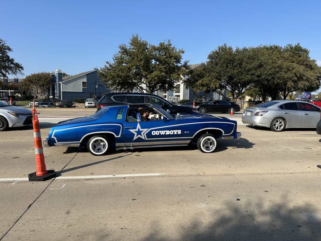 A vintage blue car painted with a large Dallas Cowboys star and logo cruises past traffic cones on a busy street, with trees and low buildings in the background.