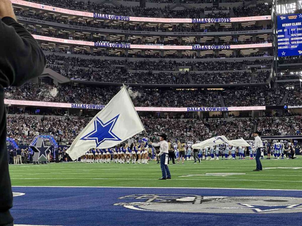 Cowboys Wrangler Flag Team members in white shirts and cowboy hats wave large white flags with blue stars across the end zone while the team and cheerleaders line up behind them.