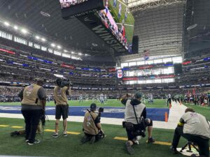 A row of NFL photographers and camera operators kneels along the goal line at AT&T Stadium, lenses trained on the action as the game unfolds under the towering roof and video board.