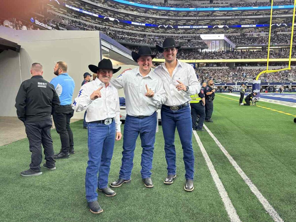 Three members of the Dallas Cowboys Wrangler Flag Team stand on the sideline at AT&T Stadium wearing white Western shirts, blue jeans, cowboy hats, and large belt buckles, smiling with the packed stands rising behind them.