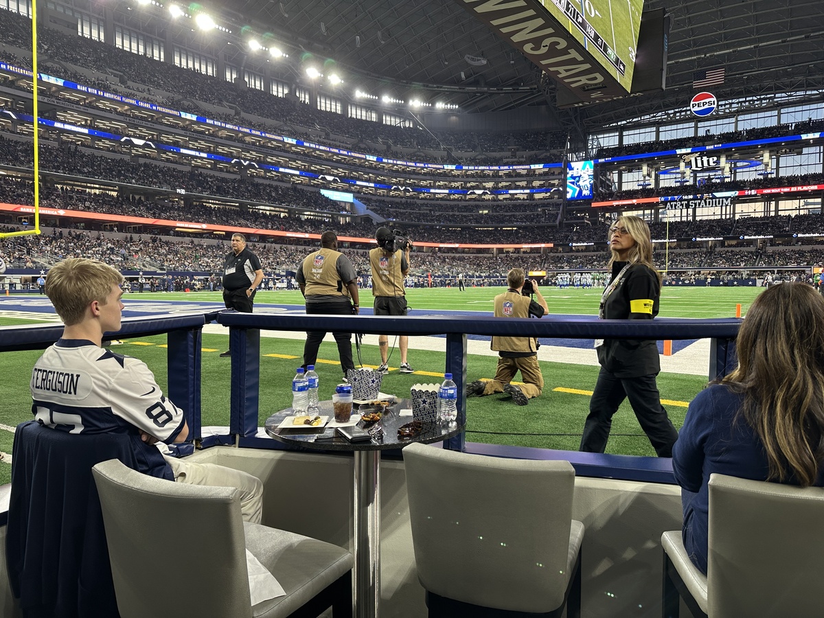 Field-level club seating at AT&T Stadium, with a young fan in a Cowboys jersey sitting at a small table covered in snacks and drinks while security staff and NFL camera crews stand just beyond the railing, watching the game unfold on the brightly lit field.