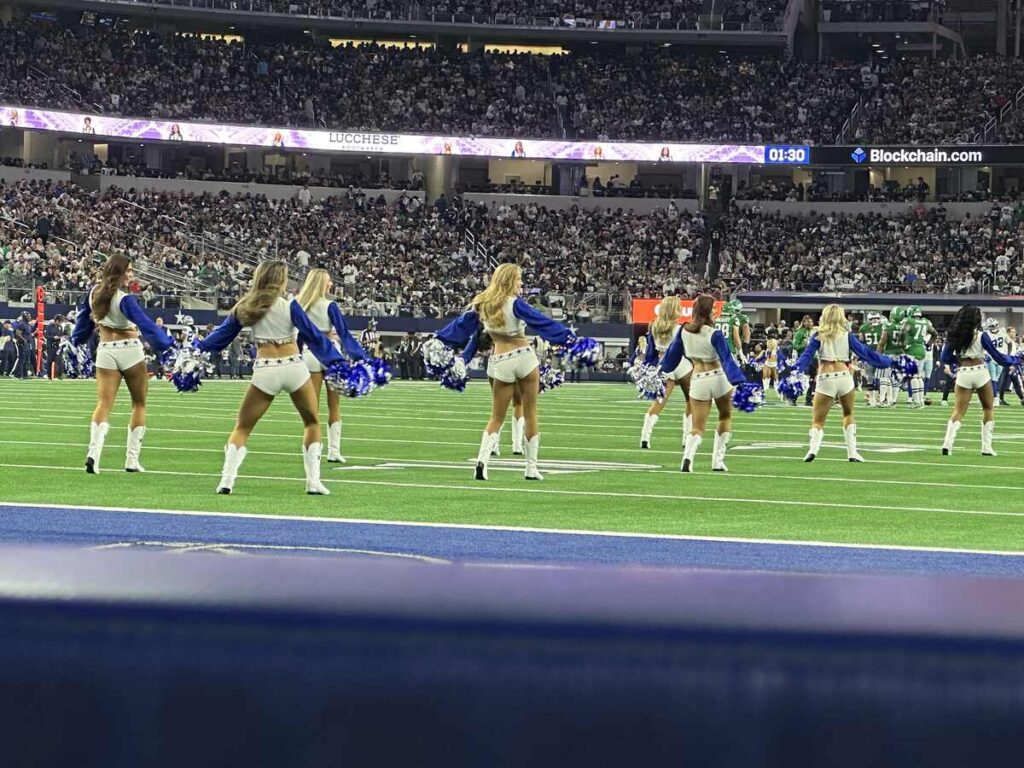 A line of Dallas Cowboys Cheerleaders performs on the field in their iconic blue-and-white uniforms and white boots, pom-poms in hand, with a full stadium of fans in the background.