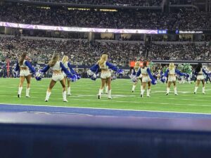 A line of Dallas Cowboys Cheerleaders performs on the field in their iconic blue-and-white uniforms and white boots, pom-poms in hand, with a full stadium of fans in the background.