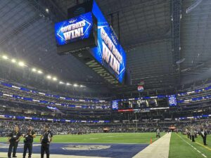 Inside AT&T Stadium, the giant centre-hung video board displays a bright blue “Cowboys Win!” graphic while security staff and lingering fans stand near the end zone.