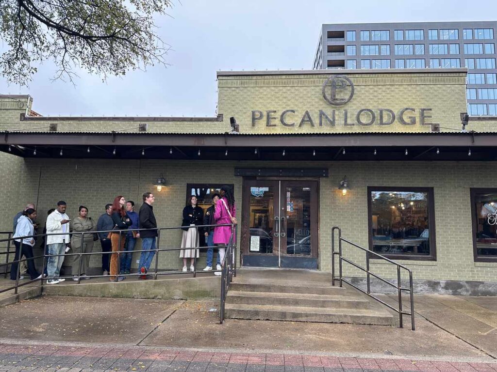 People waiting in line outside Pecan Lodge, a brick barbecue restaurant in Deep Ellum, Dallas, on an overcast day.