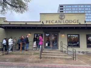 People waiting in line outside Pecan Lodge, a brick barbecue restaurant in Deep Ellum, Dallas, on an overcast day.