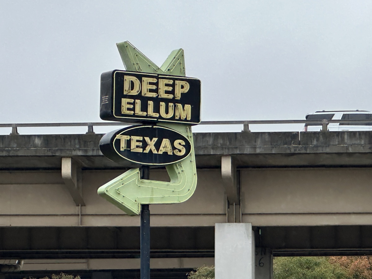 Vintage-style roadside sign reading “Deep Ellum Texas” standing in front of an elevated highway.