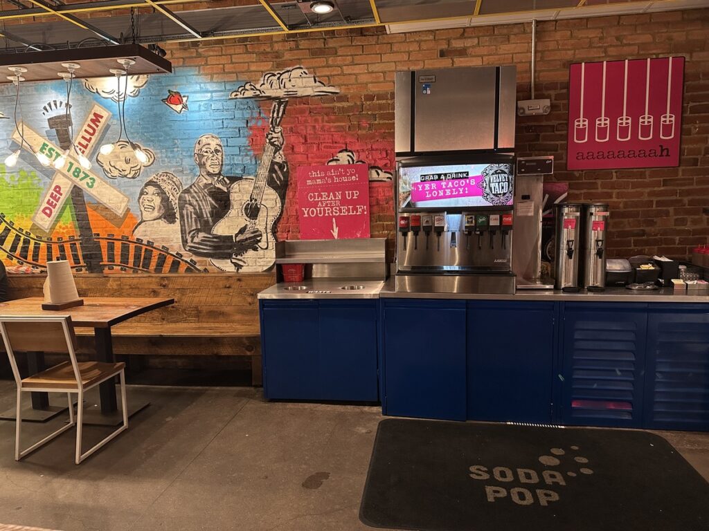 Interior of Velvet Taco in Deep Ellum with a drinks station, blue cabinets and a mural of musicians and a “Deep Ellum Est. 1873” sign on exposed brick.
