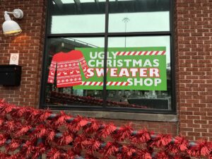 Green and red “Ugly Christmas Sweater Shop” sign in a window, framed by red tinsel garland on a brick wall.