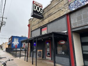 Entrance to Angry Dog bar and grill on Commerce Street in Deep Ellum, with neon “open” sign and a blue building visible further down the sidewalk.