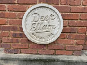 Round stone plaque reading “Deep Ellum Dallas, TX” mounted on a red brick wall.