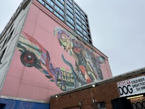 Colourful mural on a tall pink building in Deep Ellum, with musicians, abstract shapes and “Deep Ellum Texas” text above the roofline of Angry Dog.