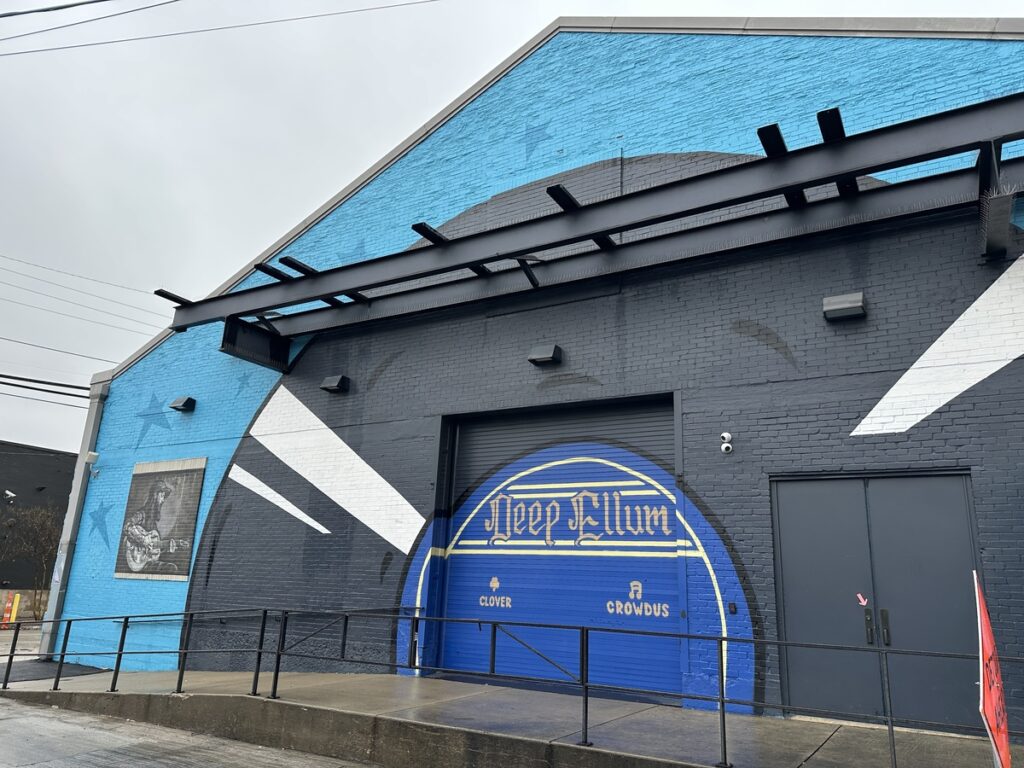 Street view of a blue and charcoal-grey warehouse-style building in Deep Ellum with a large “Deep Ellum” graphic painted on a roller door.