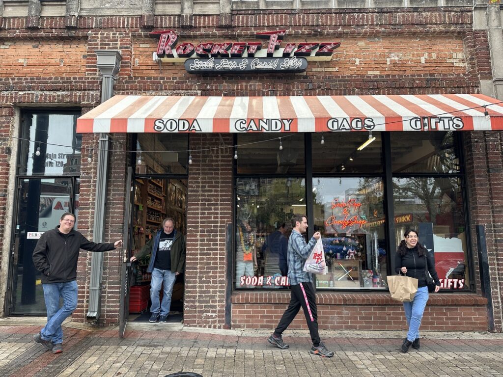 Exterior of Rocket Fizz soda and candy shop in Deep Ellum, with a red-and-white striped awning and people coming and going along the sidewalk.