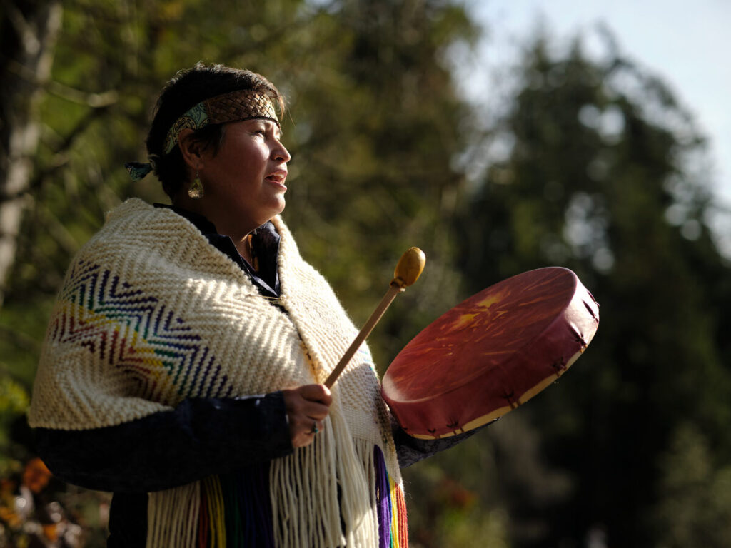 An Indigenous woman stands outdoors in soft sunlight, holding a hand drum and drumstick. She wears a woven headband and a cream-coloured shawl with colourful geometric patterns and long fringe. She looks into the distance with a calm, focused expression. Forest trees are softly blurred behind her.
