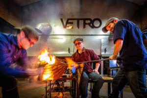 Three glassblowers work together in the Vetro Glassblowing Studio, shaping molten glass as bright flames and sparks fly. One artist sits at the bench turning the blowpipe while the others lean in to heat and sculpt the glowing piece under the industrial “VETRO” sign.