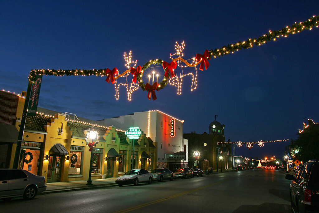 Grapevine’s Main Street at dusk, with reindeer and wreath light displays strung over the road, shopfronts glowing, and the Palace Theatre marquee lit up in the distance.