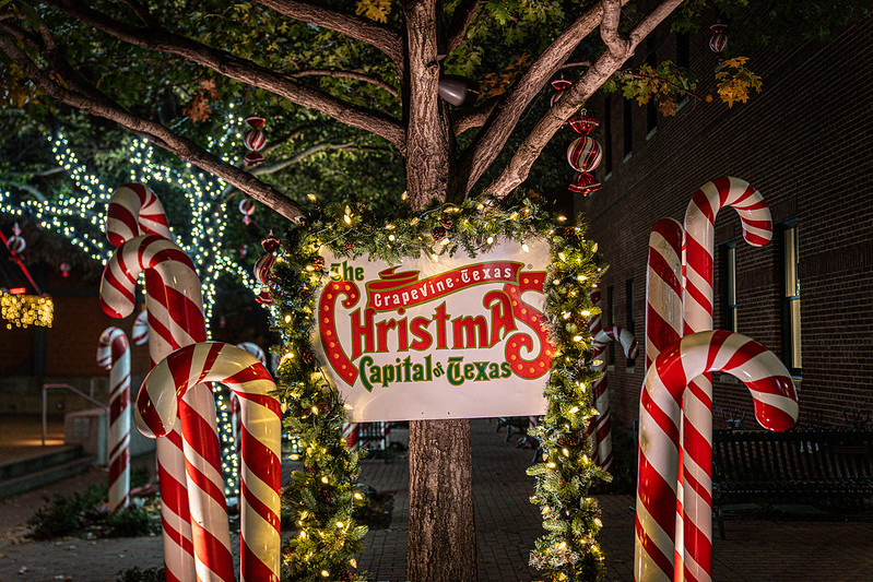 A festive sign reading “Grapevine, Texas – The Christmas Capital of Texas” framed by a lit garland, surrounded by oversized candy cane decorations and twinkling lights in the trees.