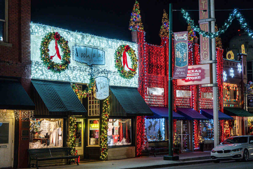 A row of historic Grapevine storefronts at night, wrapped in thousands of white and red Christmas lights with giant wreaths, striped awnings, and shop windows glowing with holiday displays.