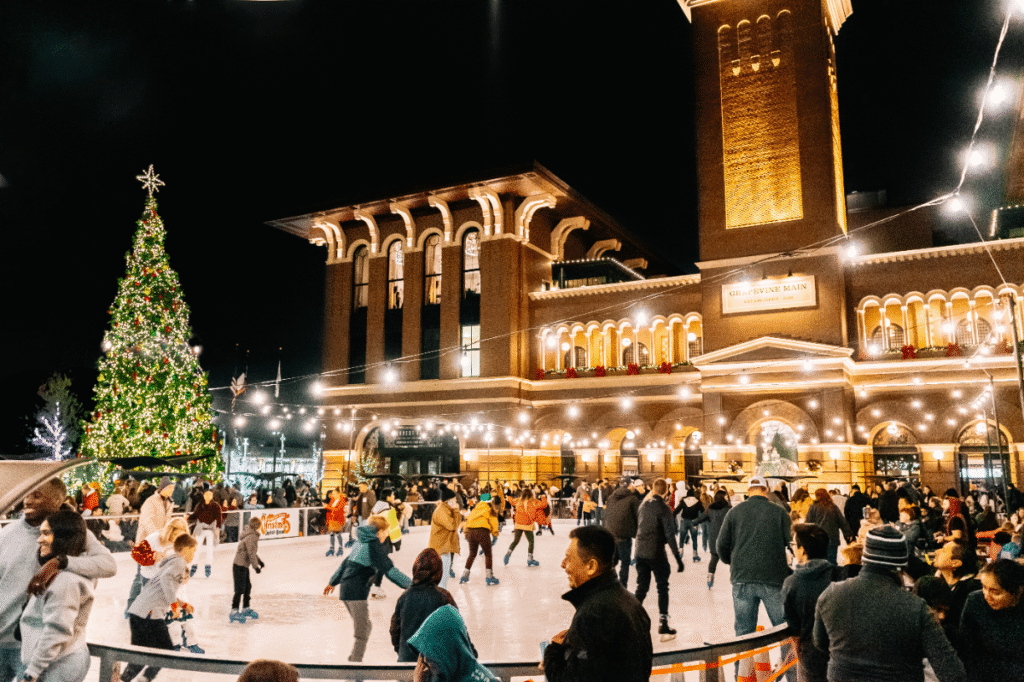 An outdoor ice rink at Peace Plaza in front of Grapevine Main Station is filled with skaters of all ages gliding under strings of warm lights. A towering, glowing Christmas tree anchors the scene, while the grand brick station building shines in the background against the night sky.