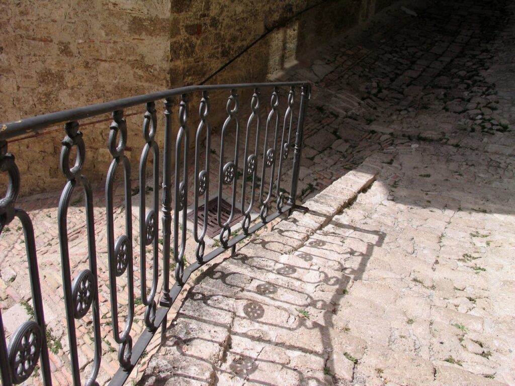 A narrow stone alley with an old iron railing casting patterned shadows, quietly suggesting barriers, thresholds, and unseen paths.