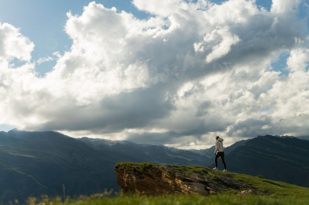 A solitary hiker stands on a grassy cliff edge beneath towering clouds and distant mountains, small against an immense landscape.